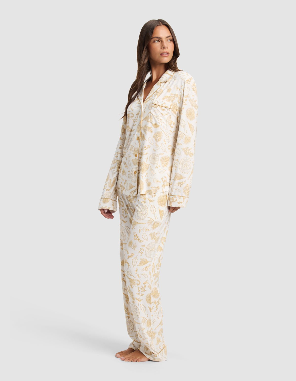A woman stands barefoot in Cozy Earth's Women's Bamboo Stretch-Knit Long Sleeve Pajama Set, featuring a white base with a gold floral print, posing against a plain light gray background.|Color:Winter Toile Dijon