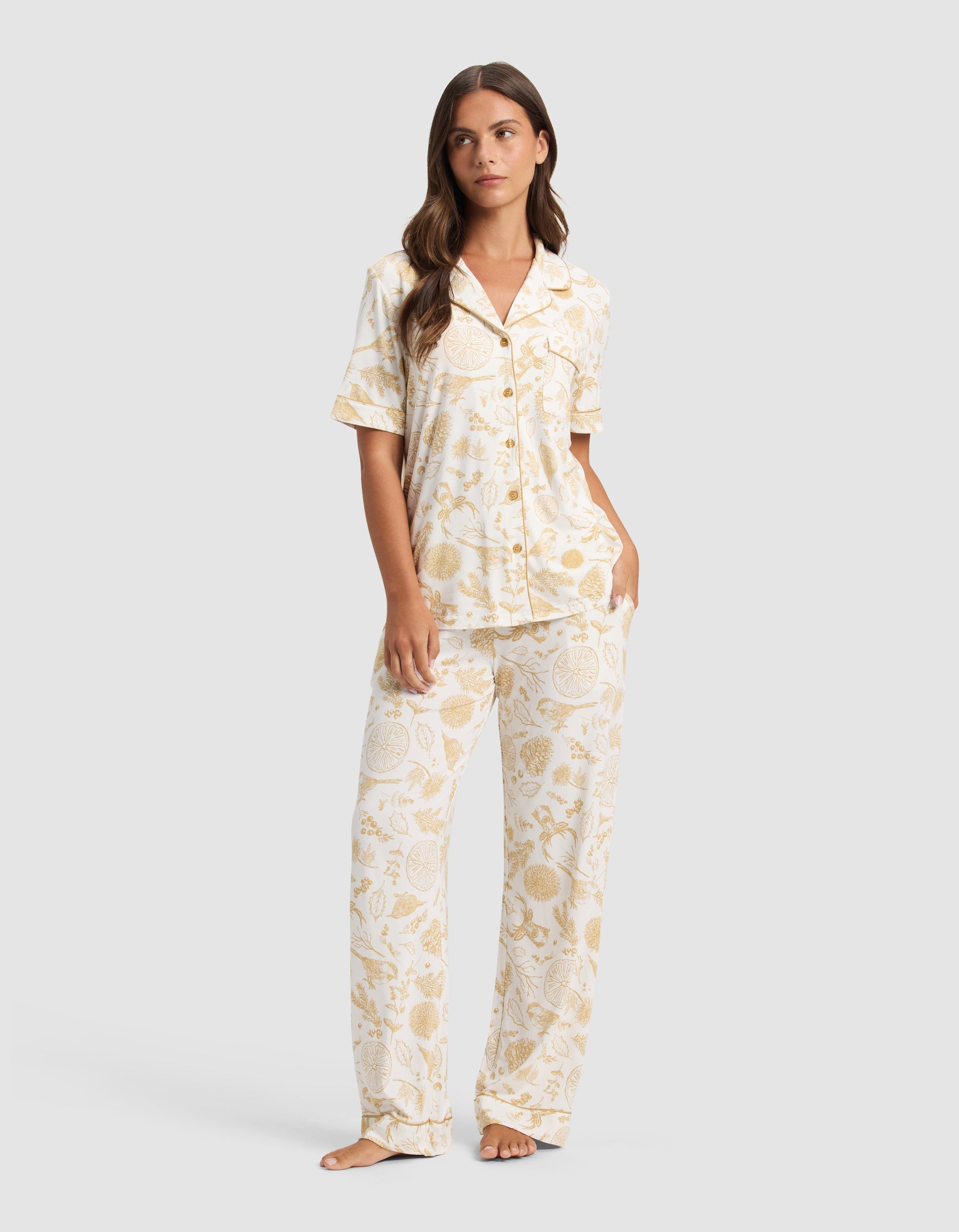 A woman with long brown hair stands barefoot, wearing Cozy Earth's Women's Bamboo Stretch-Knit Short Sleeve Pajama Set in a light yellow and white botanical print. She looks slightly to the side.|Color:Winter Toile Dijon