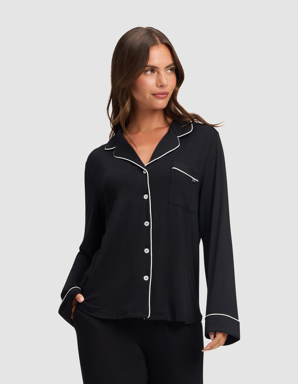 A woman models the HIDE Women's Bamboo Stretch-Knit Long Sleeve Pajama Top in black with white piping on the collar, cuffs, and pocket edges, standing against a light gray background and smiling softly. |Color:Black