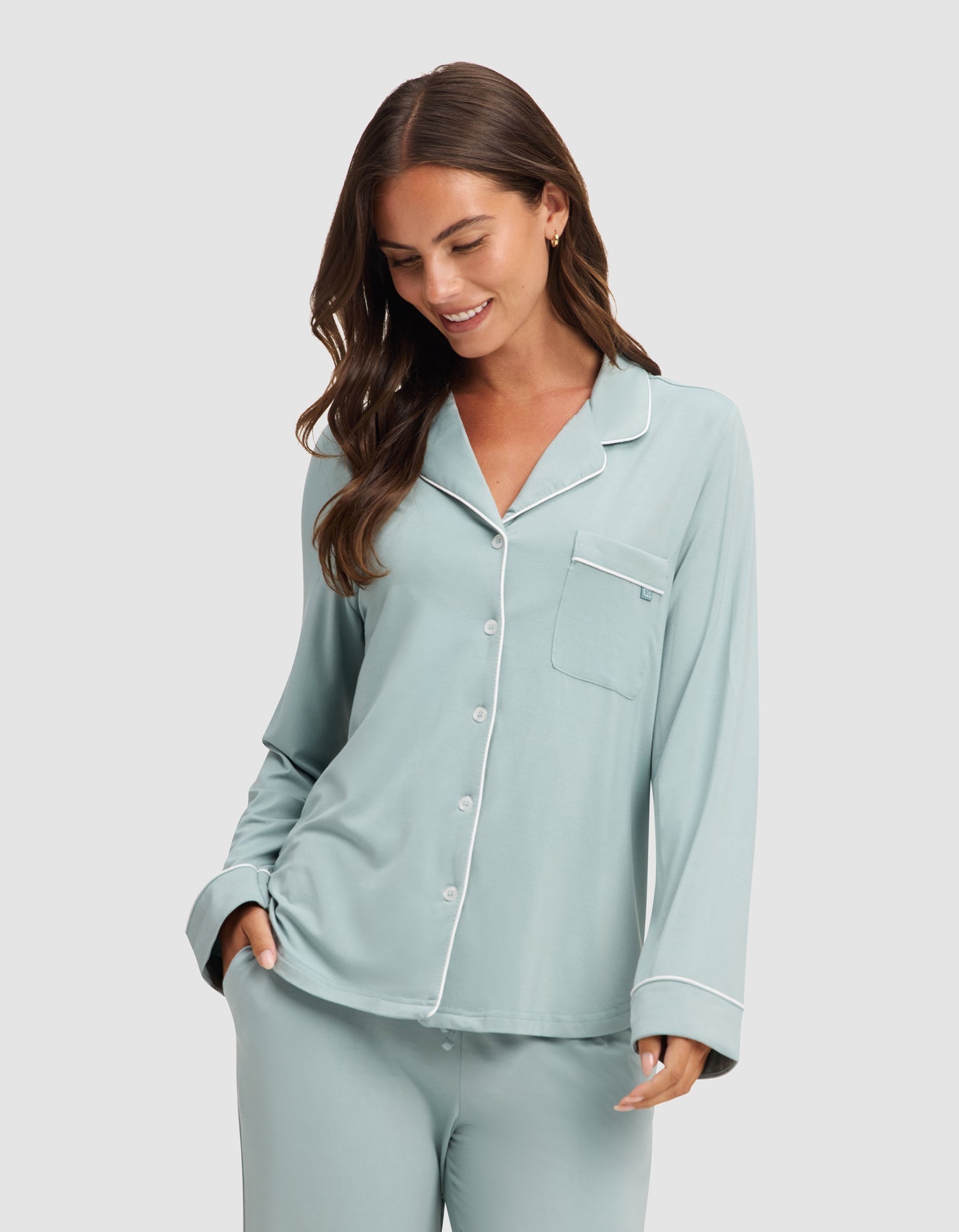 A woman smiles and looks down with one hand in her pocket, wearing HIDE's Women's Bamboo Stretch-Knit Long Sleeve Pajama Top in light blue with white piping and buttons. |Color:Brook