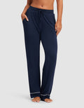 A woman wears HIDE Women's Bamboo Stretch-Knit Pajama Pant in navy, featuring white piping near the ankles, standing barefoot with hands in pockets against a plain light background. |Color:Navy