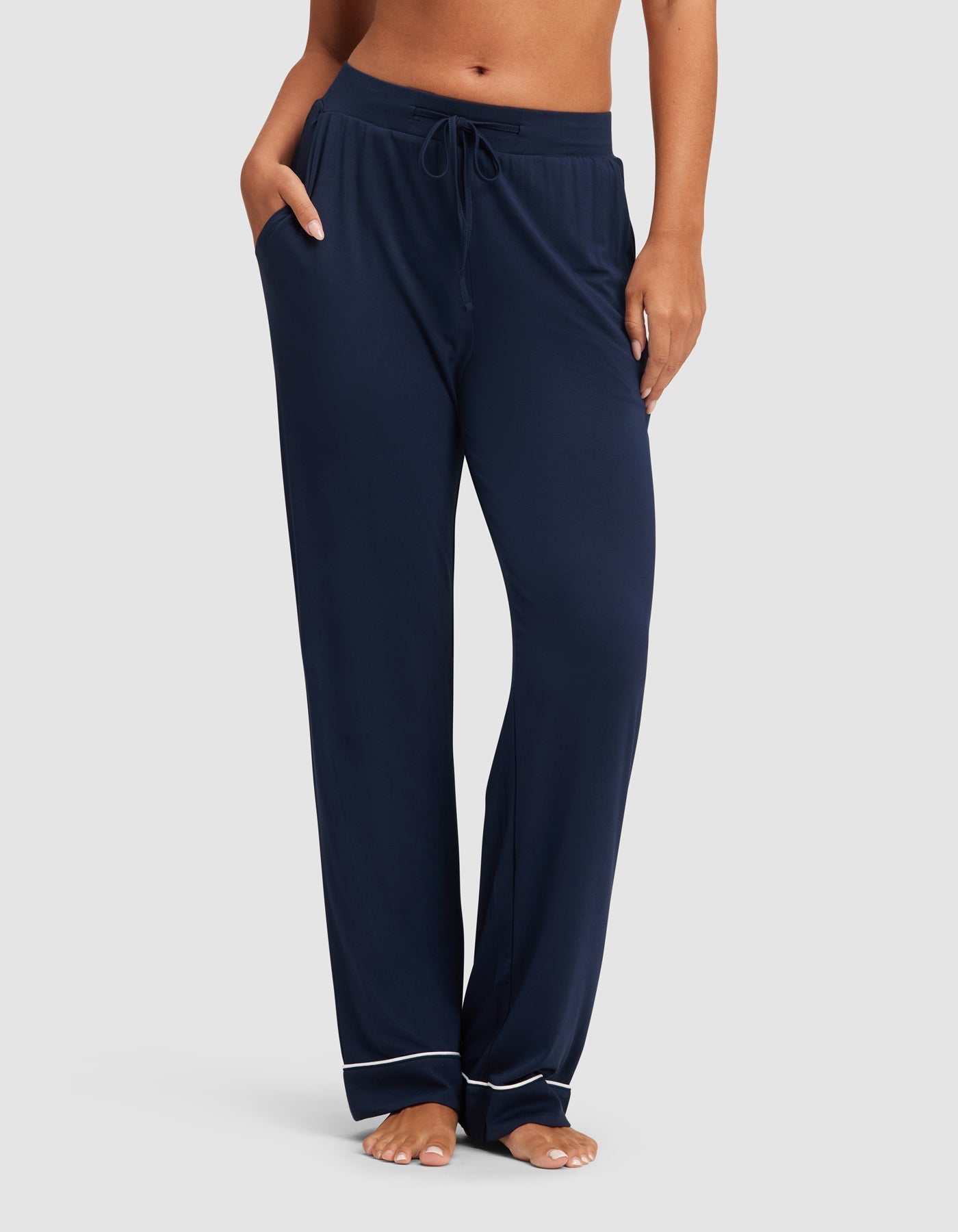 A woman wears HIDE Women's Bamboo Stretch-Knit Pajama Pant in navy, featuring white piping near the ankles, standing barefoot with hands in pockets against a plain light background. |Color:Navy