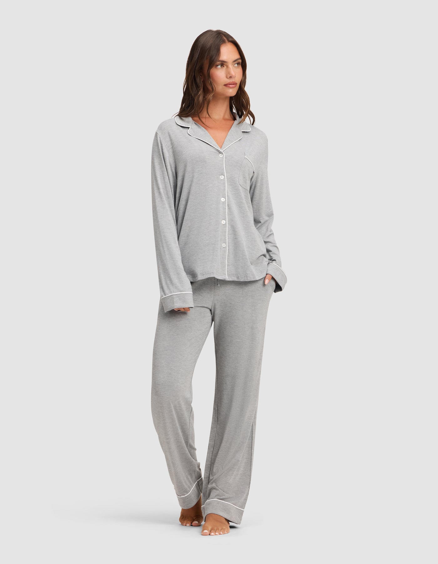 A woman stands barefoot against a plain light background, wearing the Cozy Earth Women's Bamboo Stretch-Knit Long Sleeve Pajama Set in light grey with white piping, featuring a button-up top and matching pants.  |Color:Grey