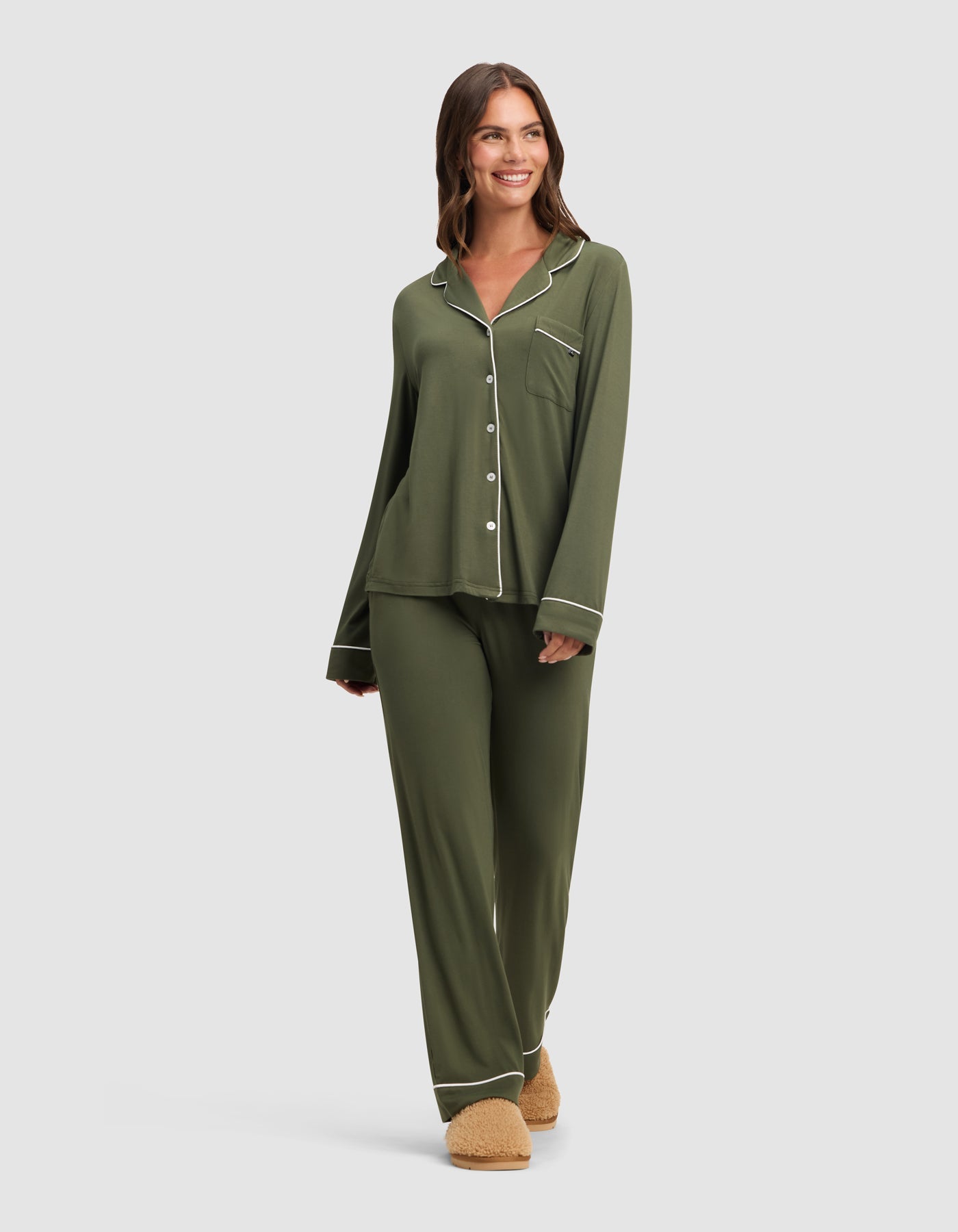 A woman smiles against a light gray background, wearing Cozy Earth's Women's Bamboo Stretch-Knit Long Sleeve Pajama Set in olive green with white piping and beige slippers.|Color:Olive