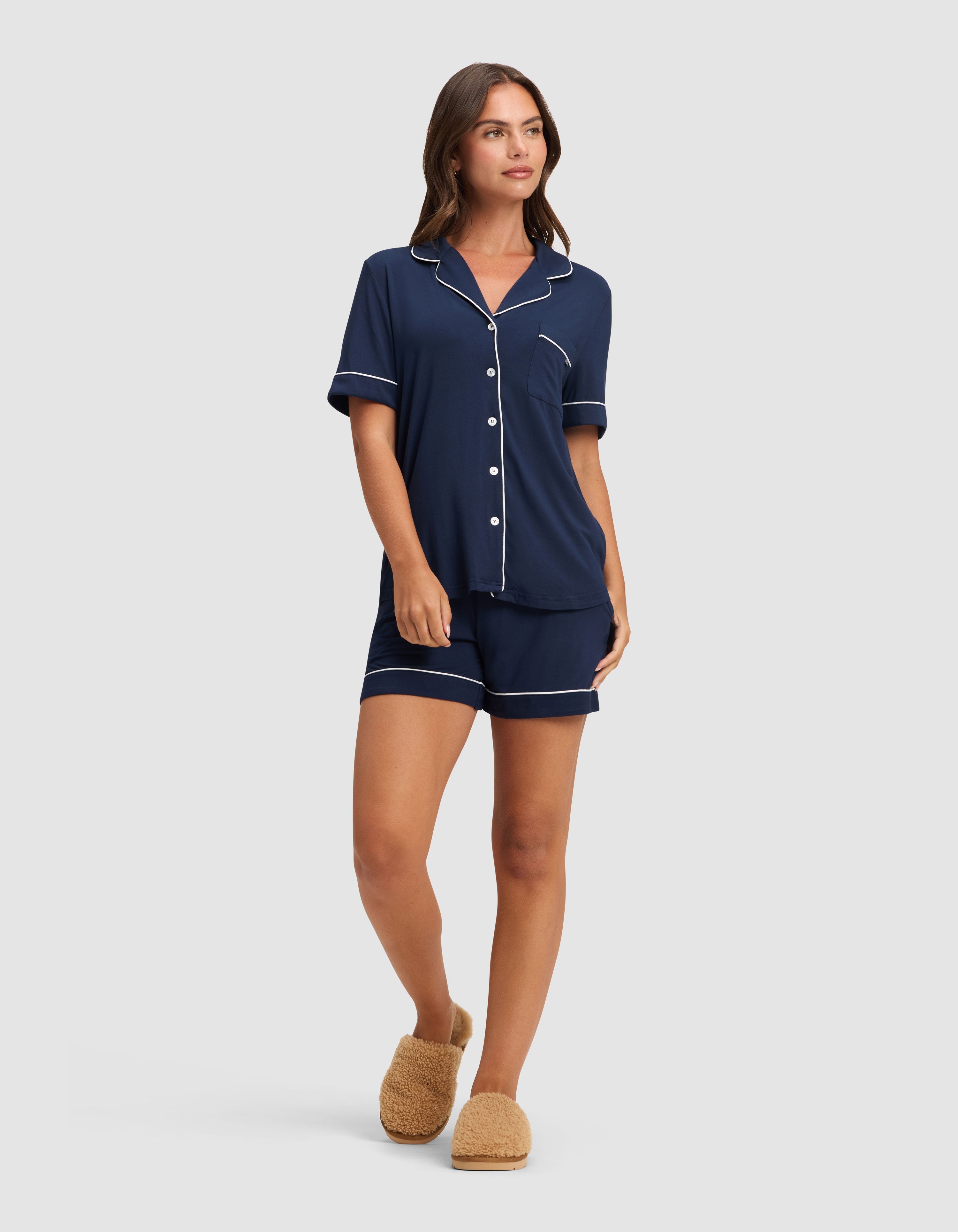 A woman wears the Cozy Earth Women's Bamboo Stretch-Knit Short Sleeve Pajama Set in navy with white piping and shorts, paired with tan fuzzy slippers, looking relaxed against a plain light background. |Color:Navy