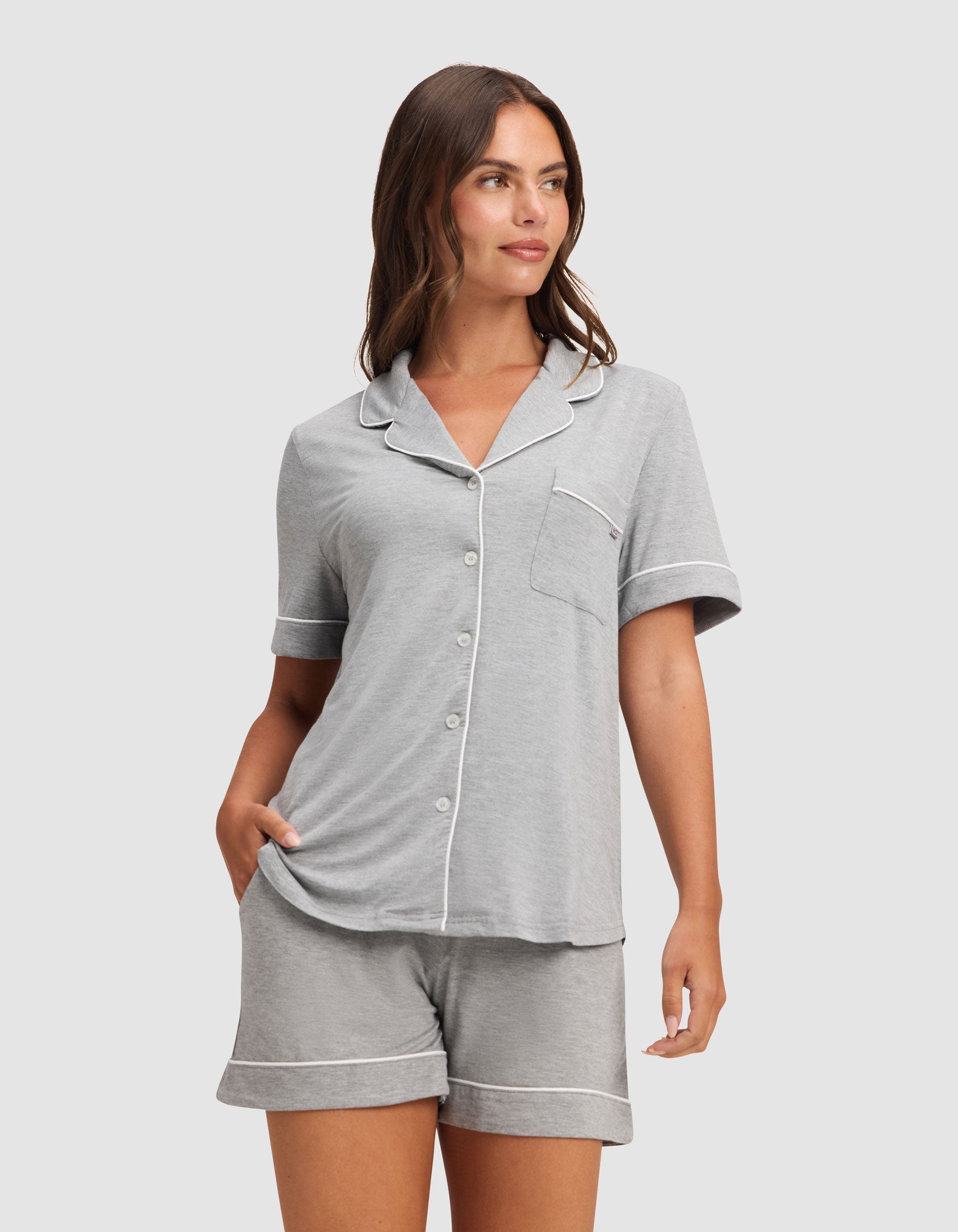 A woman models HIDE's Women's Bamboo Stretch-Knit Pajama Short, featuring a light gray short-sleeve button-up top with white piping and matching shorts, standing with one hand in her pocket against a plain background. |Color:Grey
