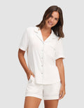 A woman with long brown hair smiles in HIDE's Women's Bamboo Stretch-Knit Pajama Short set—featuring a white short-sleeve button-up top with chest pocket and matching shorts—standing against a light gray background, one hand in her pocket. |Color:Ivory