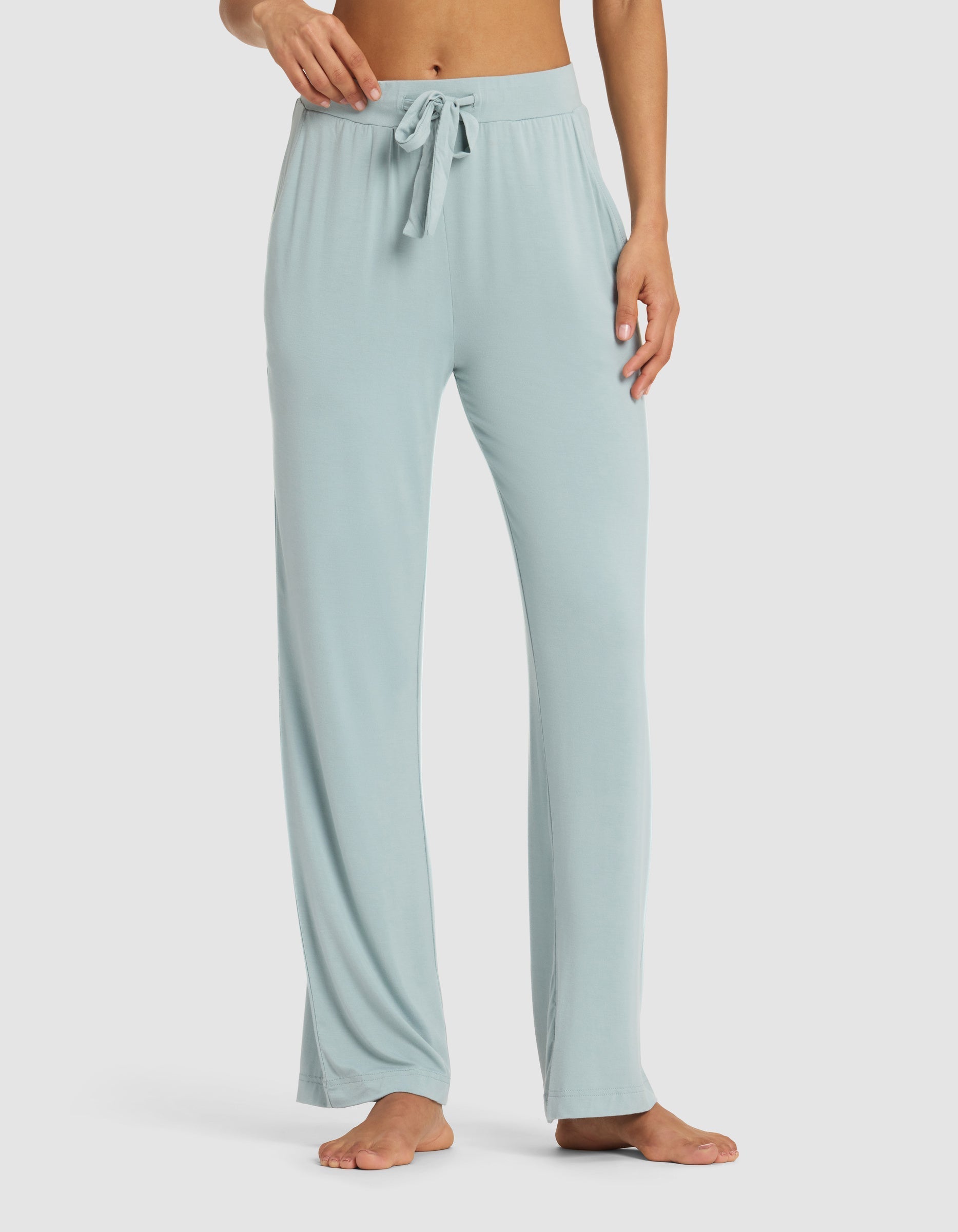 A woman models Cozy Earth’s Women's Bamboo Stretch-Knit Pant, featuring a relaxed fit, drawstring waist, and light blue color. She stands barefoot on a plain backdrop, the floor-length pants draping comfortably.|Color:Brook