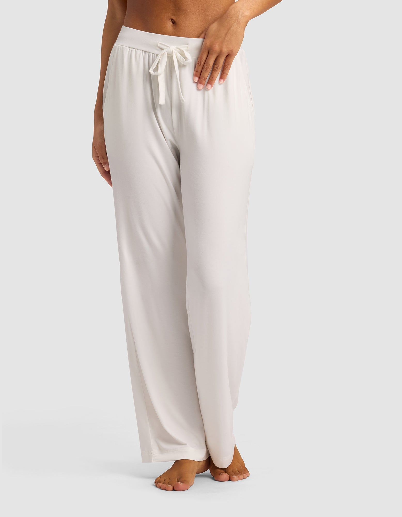 A woman stands barefoot against a plain light background, modeling the lower half of Cozy Earth's Women's Bamboo Stretch-Knit Pant—loose-fitting, light-colored lounge pants with a drawstring waist.|Color:Ivory
