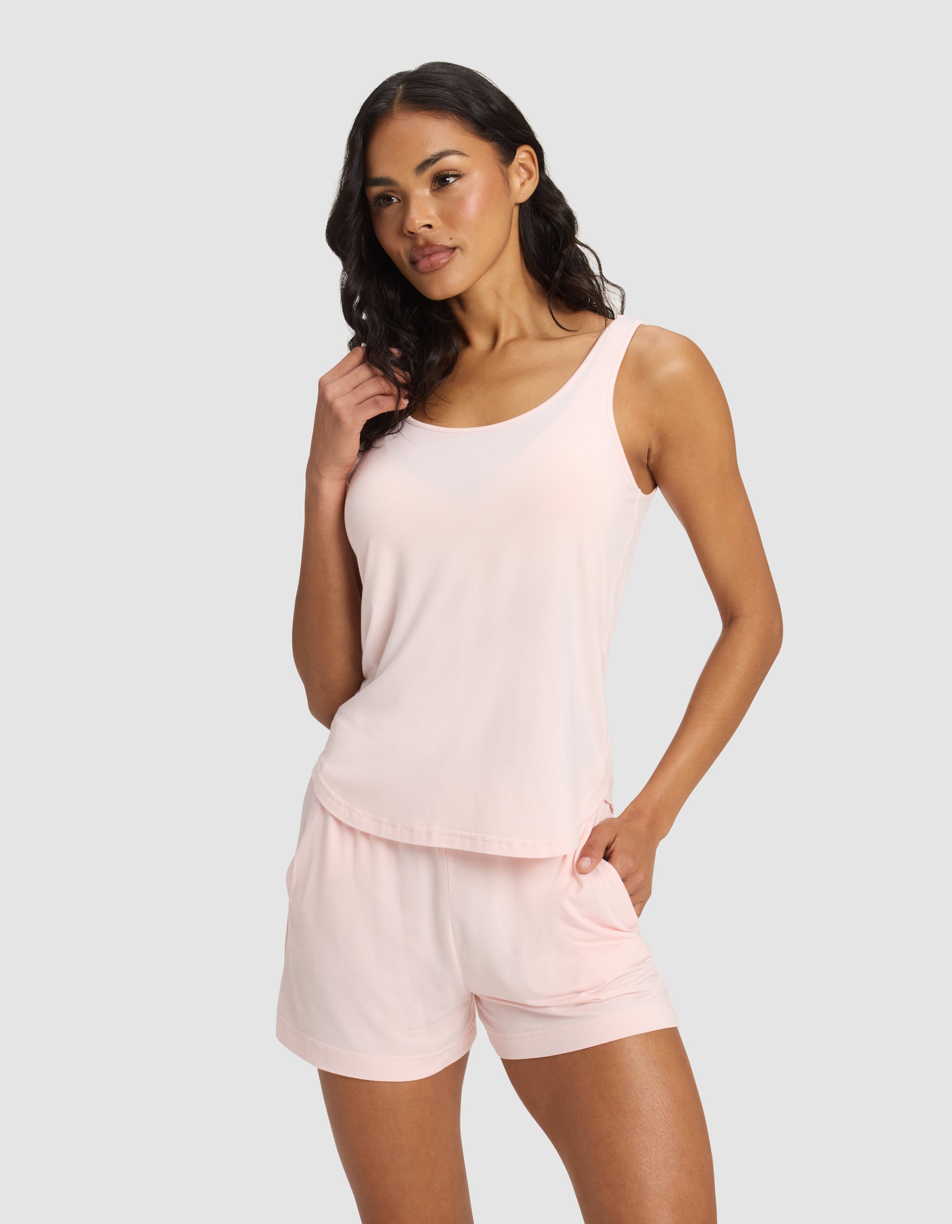 A woman with long wavy dark hair stands against a light gray background, wearing the Cozy Earth Women's Bamboo Stretch-Knit Lounge Tank in light pink and matching shorts, with one hand in her pocket, looking slightly to the side.|Color:Blossom