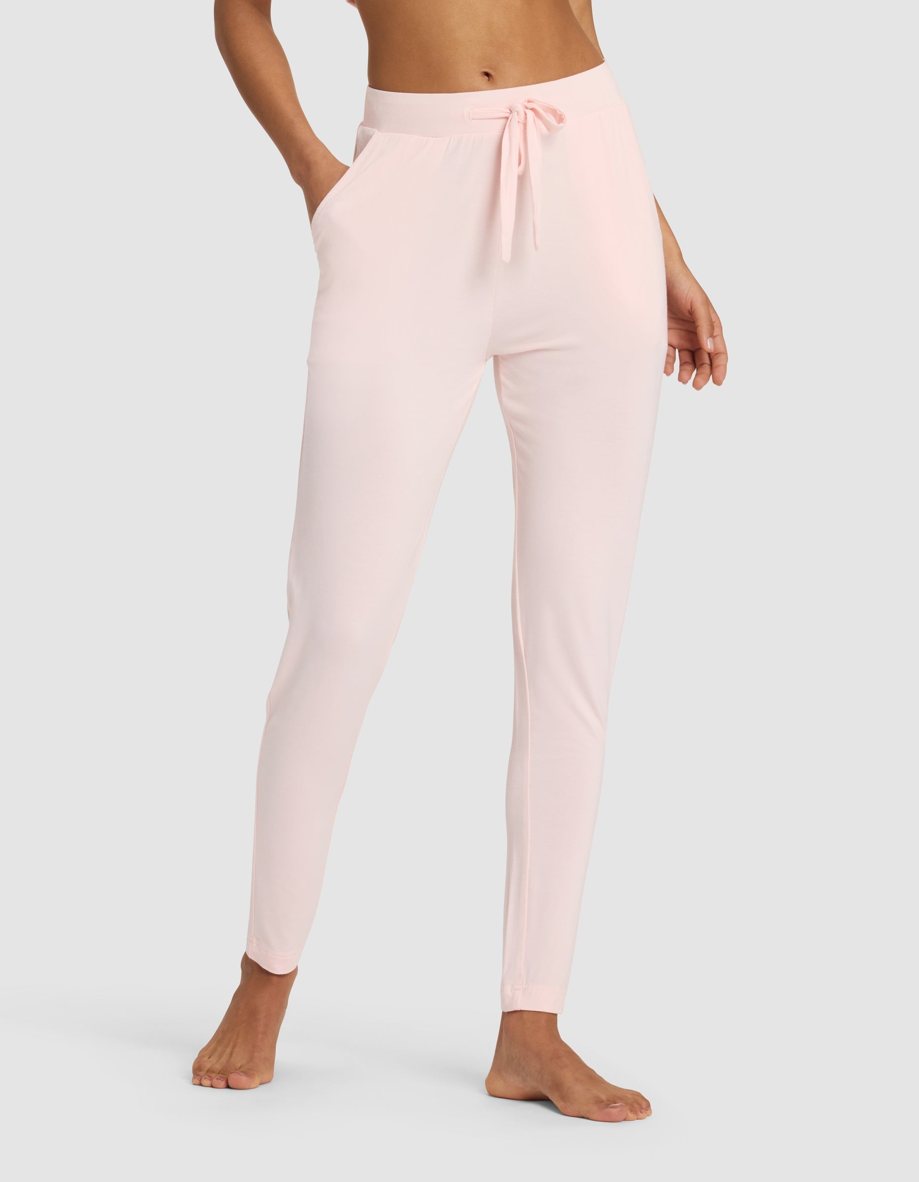 A woman wears Cozy Earth's Women's Bamboo Stretch-Knit Tapered Pant in light pink, featuring a drawstring waist and side pockets, standing barefoot against a plain light gray background.|Color:Blossom