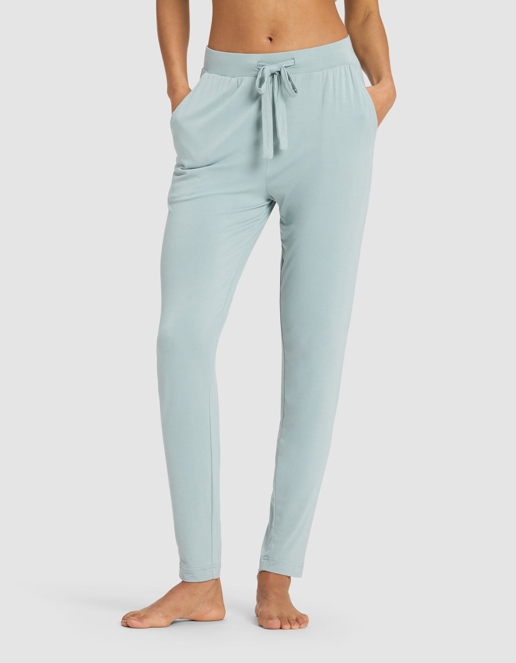 A woman models the Cozy Earth Women's Bamboo Stretch-Knit Tapered Pant in light blue, standing barefoot with hands in pockets against a plain white background.|Color:Brook